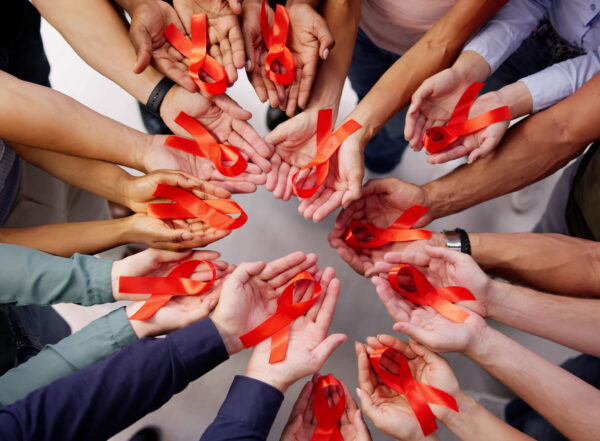 A circle of hands, all skin colors, holding red ribbons symbolizing HIV/AIDS