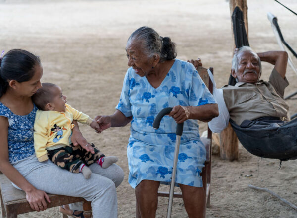 Una mujer peruana mayor con un bastón sonríe a un bebé sentado en el regazo de una mujer joven. Un hombre mayor se relaja detrás de ellos.