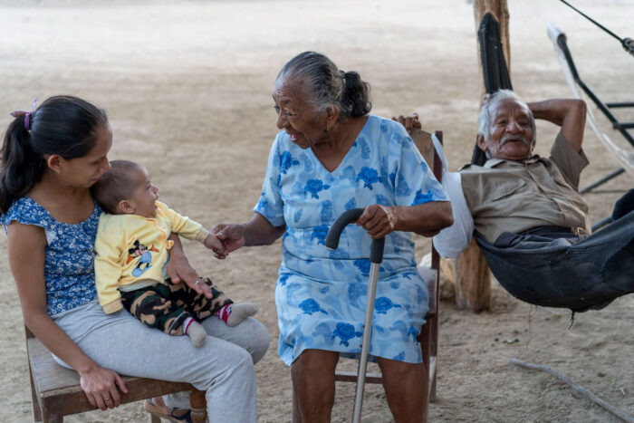 Una mujer peruana mayor con un bastón sonríe a un bebé sentado en el regazo de una mujer joven. Un hombre mayor se relaja detrás de ellos.
