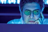 Viewed from over the top of a computer monitor, Ziyad Al-Aly, MD, is cast in indigo light. He looks intently into the monitor, facing the camera, cheek resting in his left hand. Rows of phosphor green numerals glow on his eyeglasses, reflected back from the screen before him.