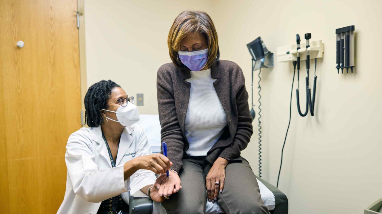 Lenise Cummings-Vaughn, MD, a Black woman, draws a stylus across the hand of Stephanie Griffin, who also is Black. The women are in a doctor's office, and Dr. Cummings-Vaughn is conducting a neurological test.
