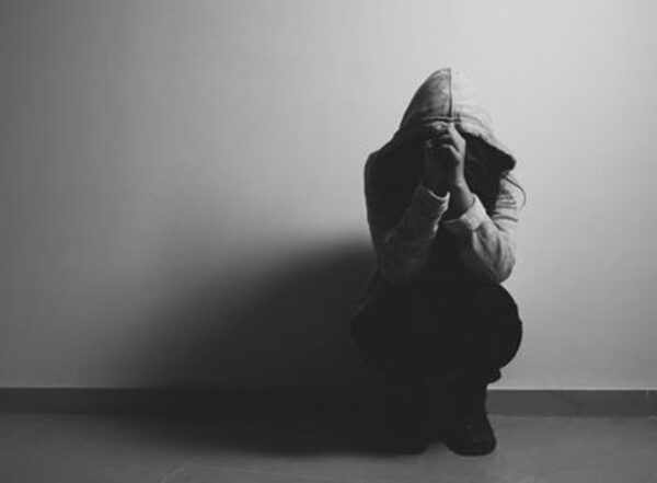 Black-and-white photo of a person wearing a hoody squatting against a wall with hands raised to face.