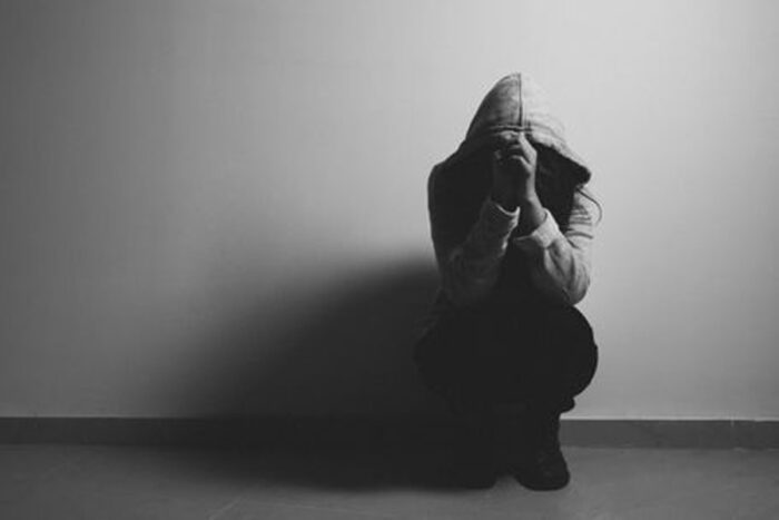 Black-and-white photo of a person wearing a hoody squatting against a wall with hands raised to face.