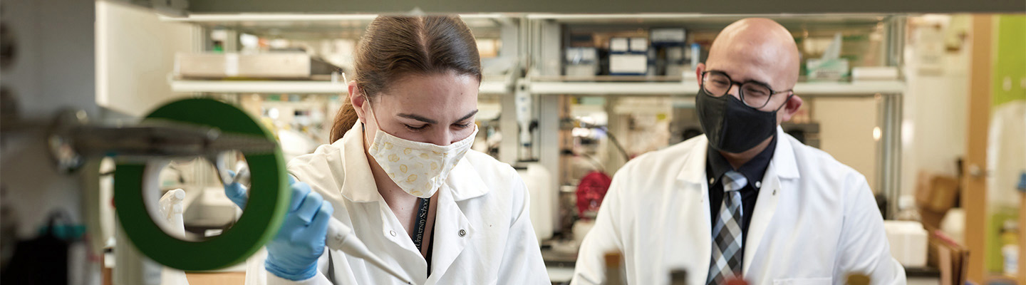 Looking between shelves of vials, we see a graduate student and faculty member white lab coats and face masks. The faculty member watches while the student adds liquid to vials using a pipette.