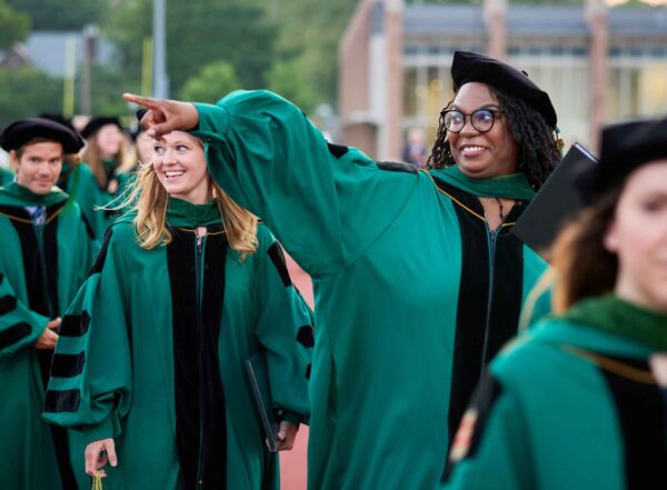 A young Black woman in emerald green regalia smiles broadly and points to an acquaintance as she walks in the Commencement processional
