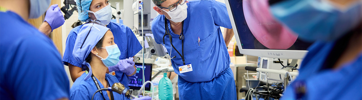 In a brightly lit operating room, a group including a medical student, resident, fellow and several physicians wear bright blue scrubs, hair covers, gloves and face masks. Everyone is leaning in, intently watching the surgery on a large monitor.