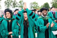 At the front of a crowd of graduates at the WashU Commencement ceremony, three graduates wearing WashU green gowns and doctoral tams and hoods stand and cheer excitedly. "Tam" is a fancy word for the velvet eight-sided hat that doctoral candidates typical wear.