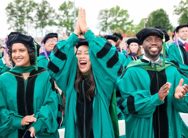 At the front of a crowd of graduates at the WashU Commencement ceremony, three graduates wearing WashU green gowns and doctoral tams and hoods stand and cheer excitedly. "Tam" is a fancy word for the velvet eight-sided hat that doctoral candidates typical wear.