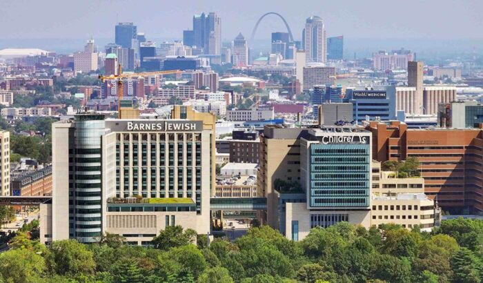 Aerial photo of the medical campus taken from above Forest Park looking east. The city stretches behind a cluster of hospital and research towers, with the Arch and downtown St. Louis distantly blue on the horizon.